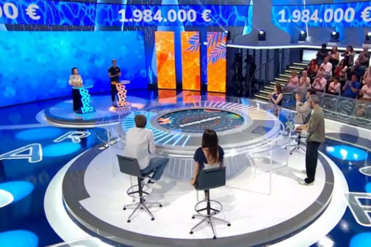 Un plató de concurs televisiu, que és 'Pasapalabra', amb dos concursants drets als faristols, dues persones assegudes d’esquena i un presentador dret, amb públic al fons i una pantalla que mostra una quantitat de diners. Un plató de concurs televisiu, que és 'Pasapalabra', amb dos concursants drets als faristols, dues persones assegudes d’esquena i un presentador dret, amb públic al fons i una pantalla que mostra una quantitat de diners.