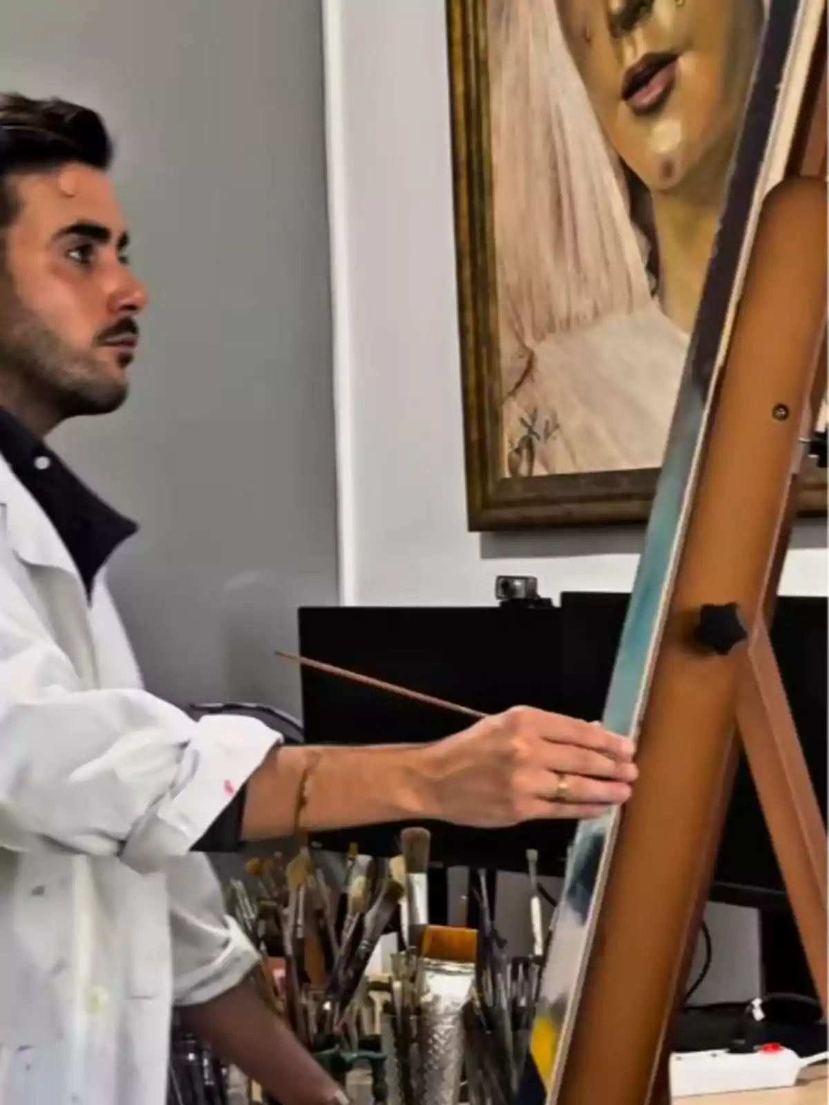 Alberto Rubio con bata blanca pintando en un caballete en un estudio de arte, rodeado de pinceles y materiales, con un cuadro de una mujer al fondo