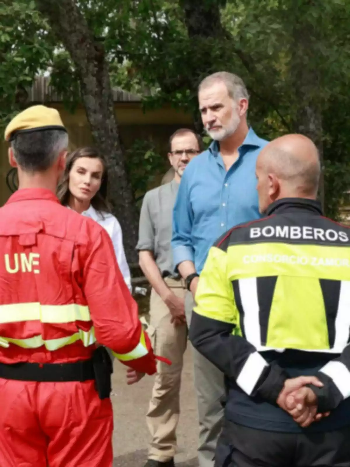 Several people are talking outdoors, including two emergency personnel in UME uniforms and firefighters, while three other people watch alertly. Several people are talking outdoors, including two emergency personnel in UME uniforms and firefighters, while three other people watch alertly.