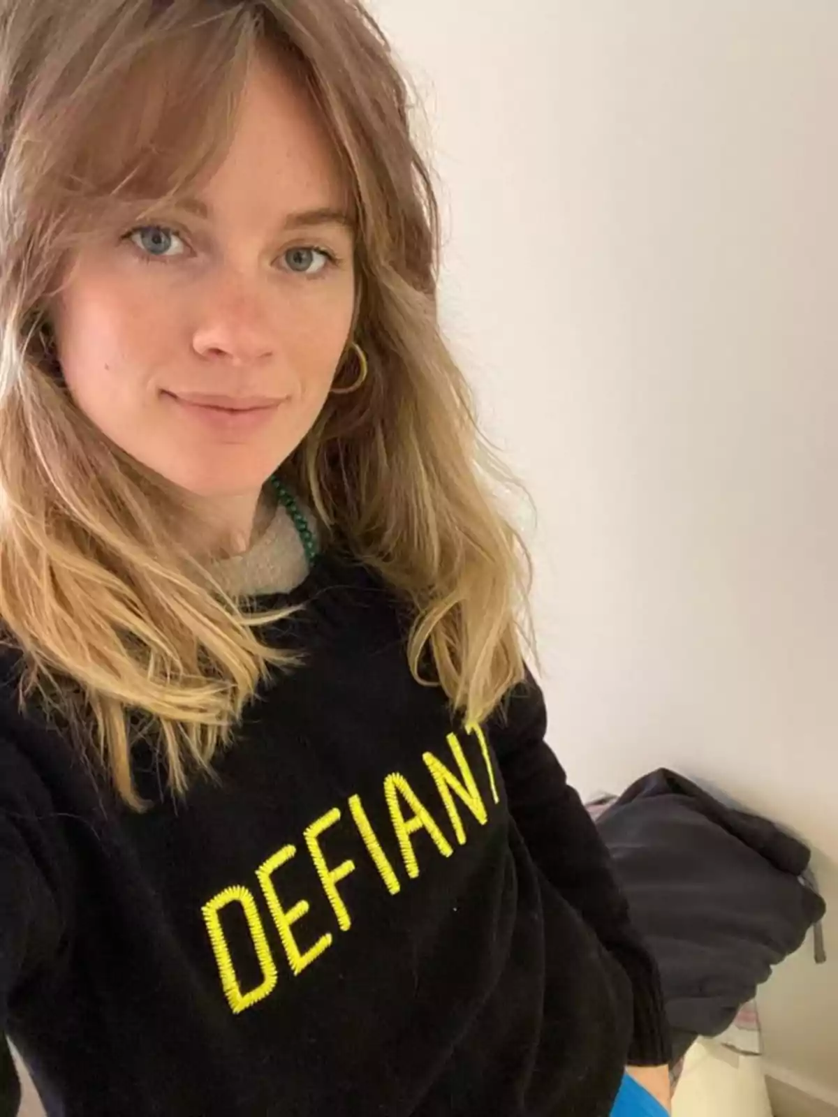 Cressida Bonas con suéter negro con la palabra DEFIANT en letras amarillas, mirando a la cámara y sonriendo levemente en un ambiente interior iluminado. Cressida Bonas con suéter negro con la palabra DEFIANT en letras amarillas, mirando a la cámara y sonriendo levemente en un ambiente interior iluminado.