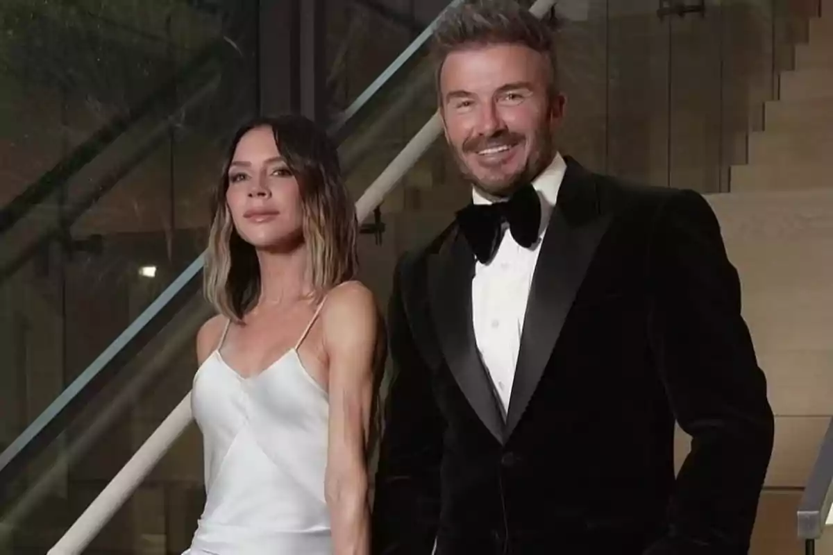 Victoria y David Beckham elegantemente vestida posa en una escalera, ella lleva un vestido blanco satinado y él un esmoquin negro con moño Victoria y David Beckham elegantemente vestida posa en una escalera, ella lleva un vestido blanco satinado y él un esmoquin negro con moño
