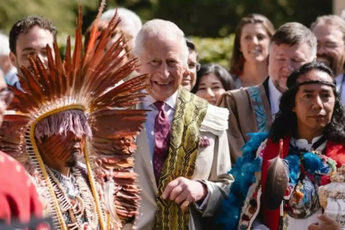 A group of people smiles outdoors, including an older man in a light suit and tie, accompanied by two men wearing traditional Indigenous clothing and headdresses made of feathers and bright colors. A group of people smiles outdoors, including an older man in a light suit and tie, accompanied by two men wearing traditional Indigenous clothing and headdresses made of feathers and bright colors.