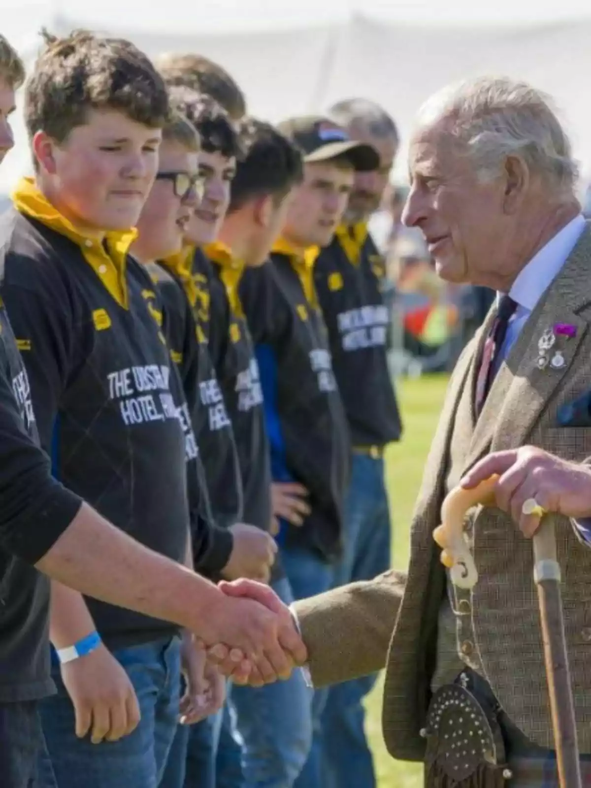 King Charles III, elegantly dressed, shakes hands with a young man while other young people watch in line at an outdoor event. King Charles III, elegantly dressed, shakes hands with a young man while other young people watch in line at an outdoor event.