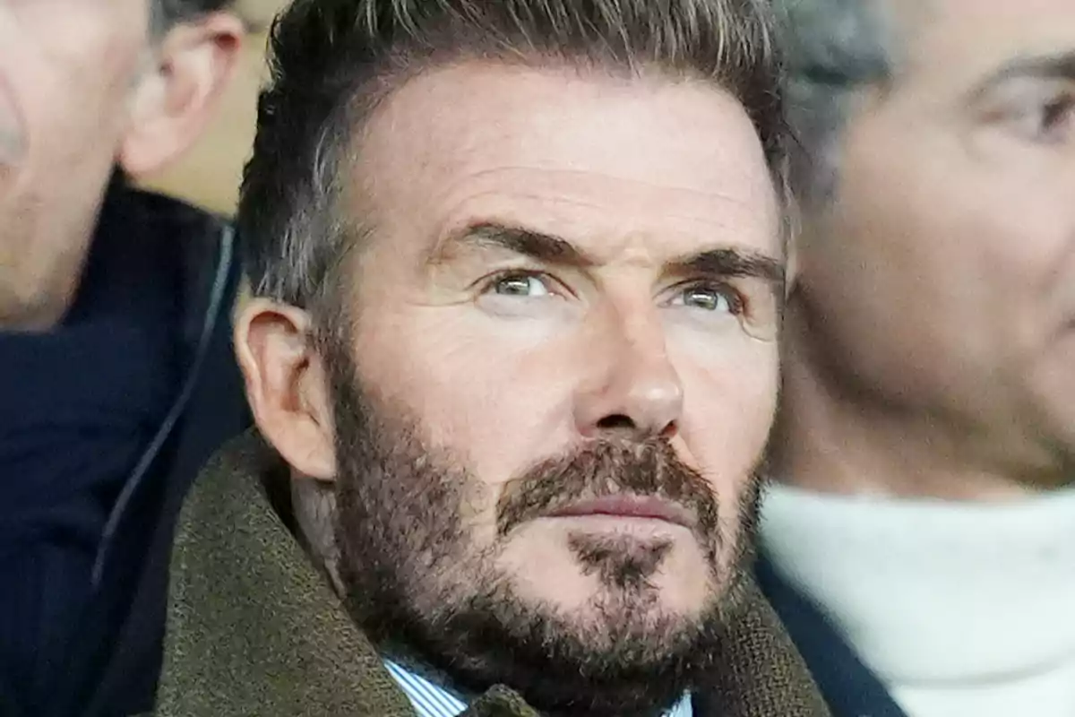 David Beckham mirando hacia el frente con expresión seria en un entorno con otras personas desenfocadas al fondo