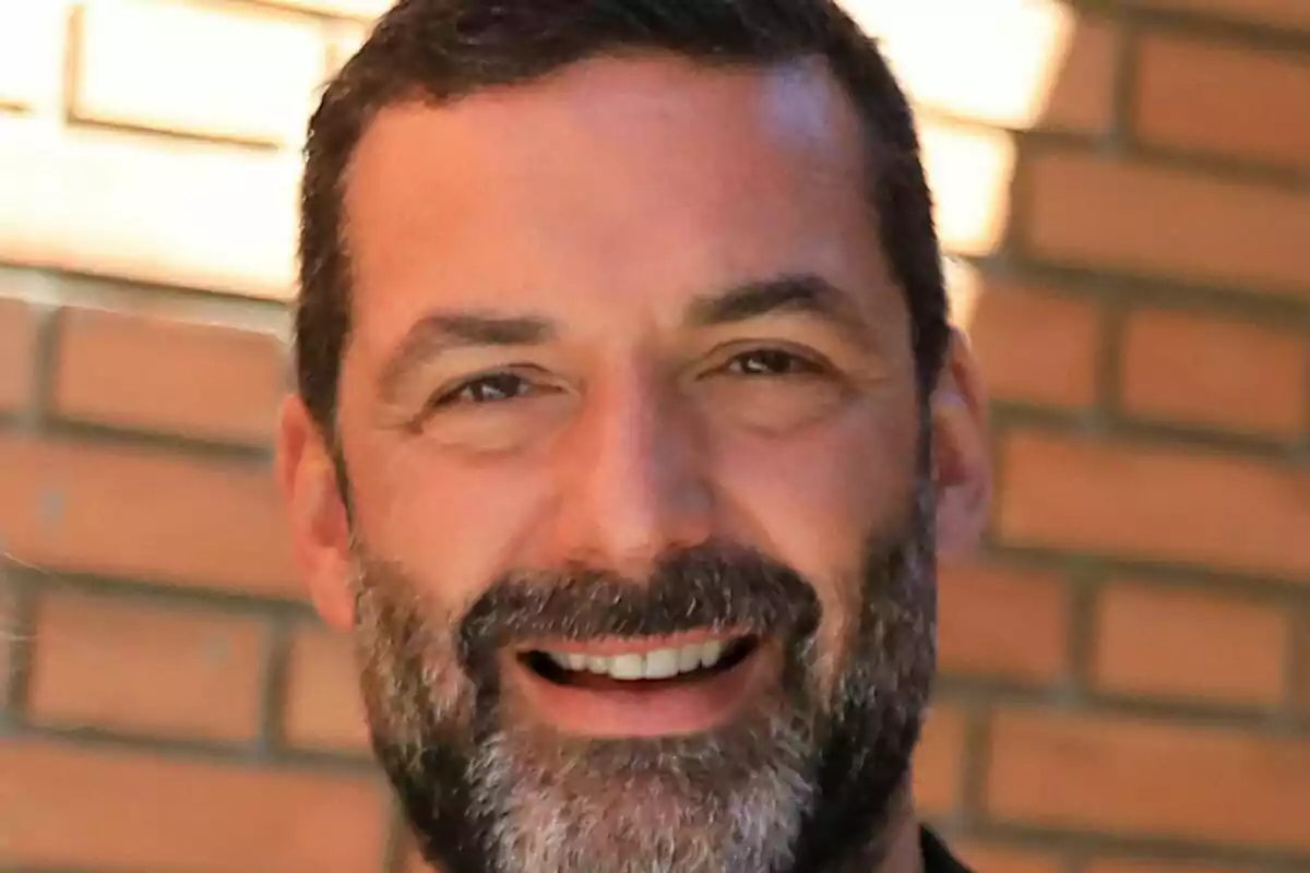 Primer plano de Jaime Anglada con barba y cabello corto sonriendo frente a una pared de ladrillos.