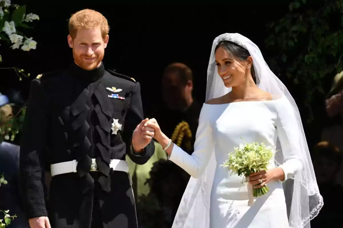 Prince Harry and Meghan Markle, dressed as bride and groom, smile and hold hands during an outdoor wedding.