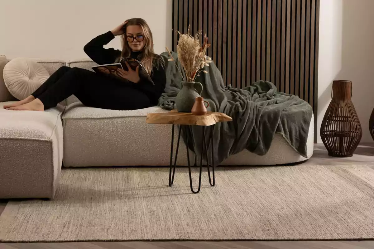 Mujer sentada en un sofá leyendo una revista en una sala decorada con tonos neutros y detalles de madera
