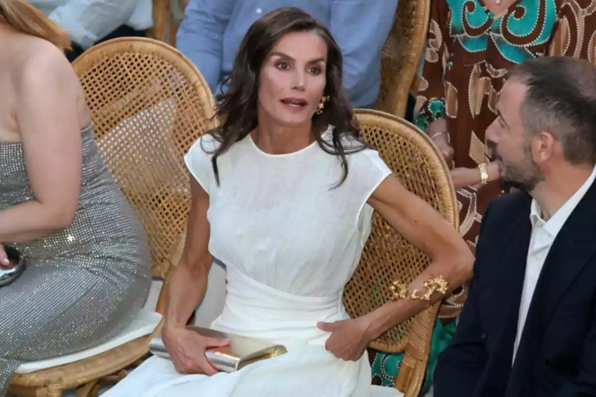 La reina Letícia amb vestit blanc asseguda en una cadira de vímet durant un esdeveniment social conversant amb un home La reina Letícia amb vestit blanc asseguda en una cadira de vímet durant un esdeveniment social conversant amb un home