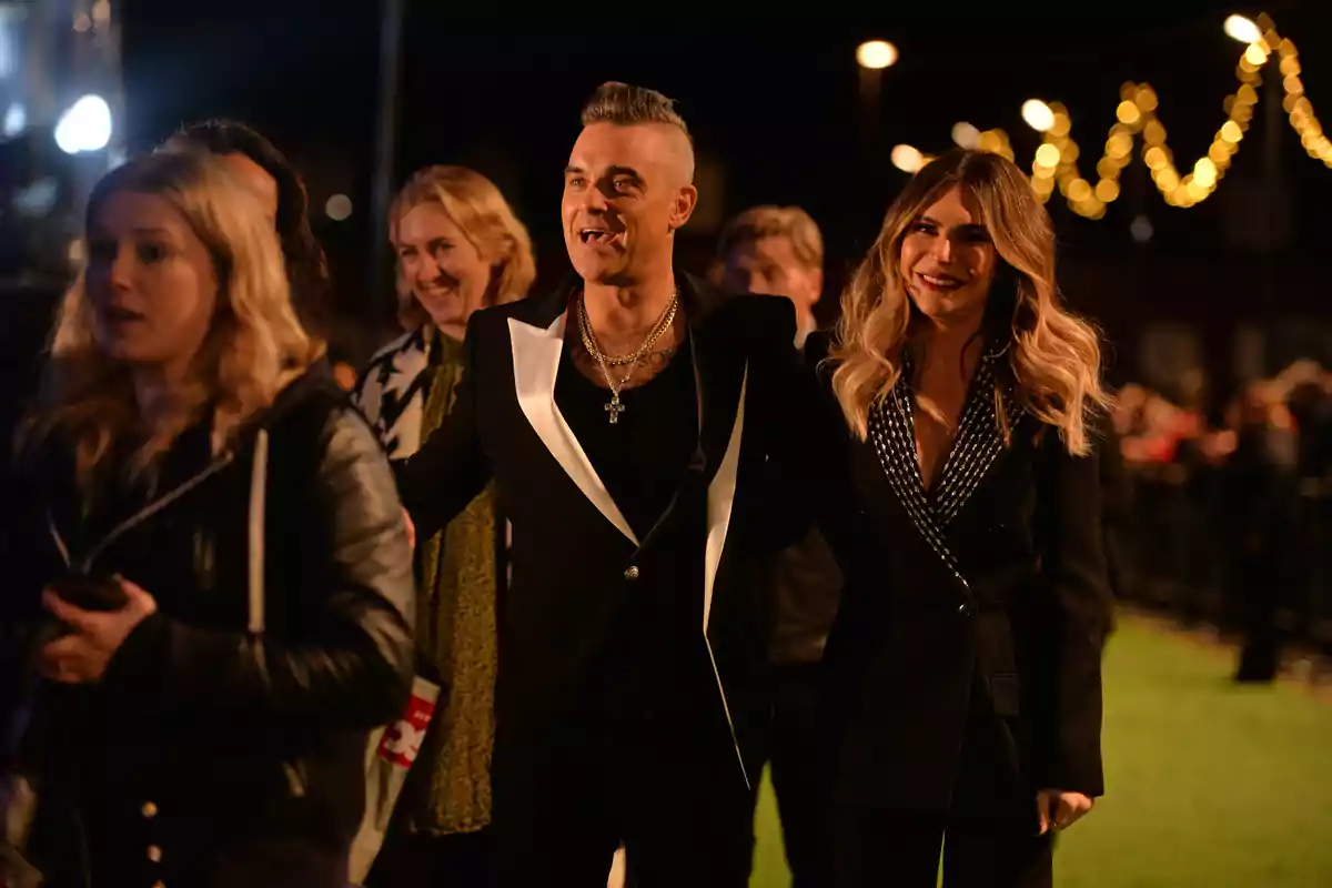 Robbie Williams and Ayda Field smile as they walk together at an outdoor nighttime event surrounded by other people and decorative lights in the background. Robbie Williams and Ayda Field smile as they walk together at an outdoor nighttime event surrounded by other people and decorative lights in the background.