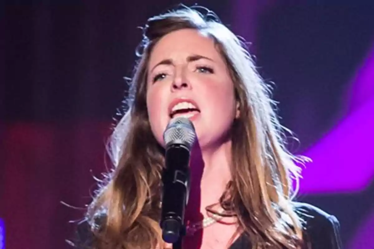 Rose Farquhar cantant en un escenari amb micròfon a la mà i llums de colors al fons Rose Farquhar cantant en un escenari amb micròfon a la mà i llums de colors al fons