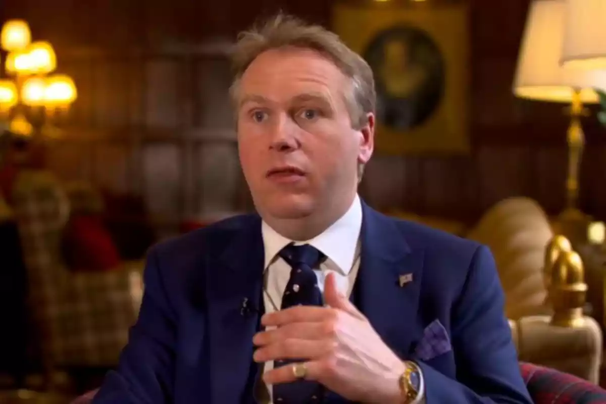 Grant Harrold amb vestit blau parlant en una sala elegantment decorada amb làmpades i mobles clàssics Grant Harrold amb vestit blau parlant en una sala elegantment decorada amb làmpades i mobles clàssics