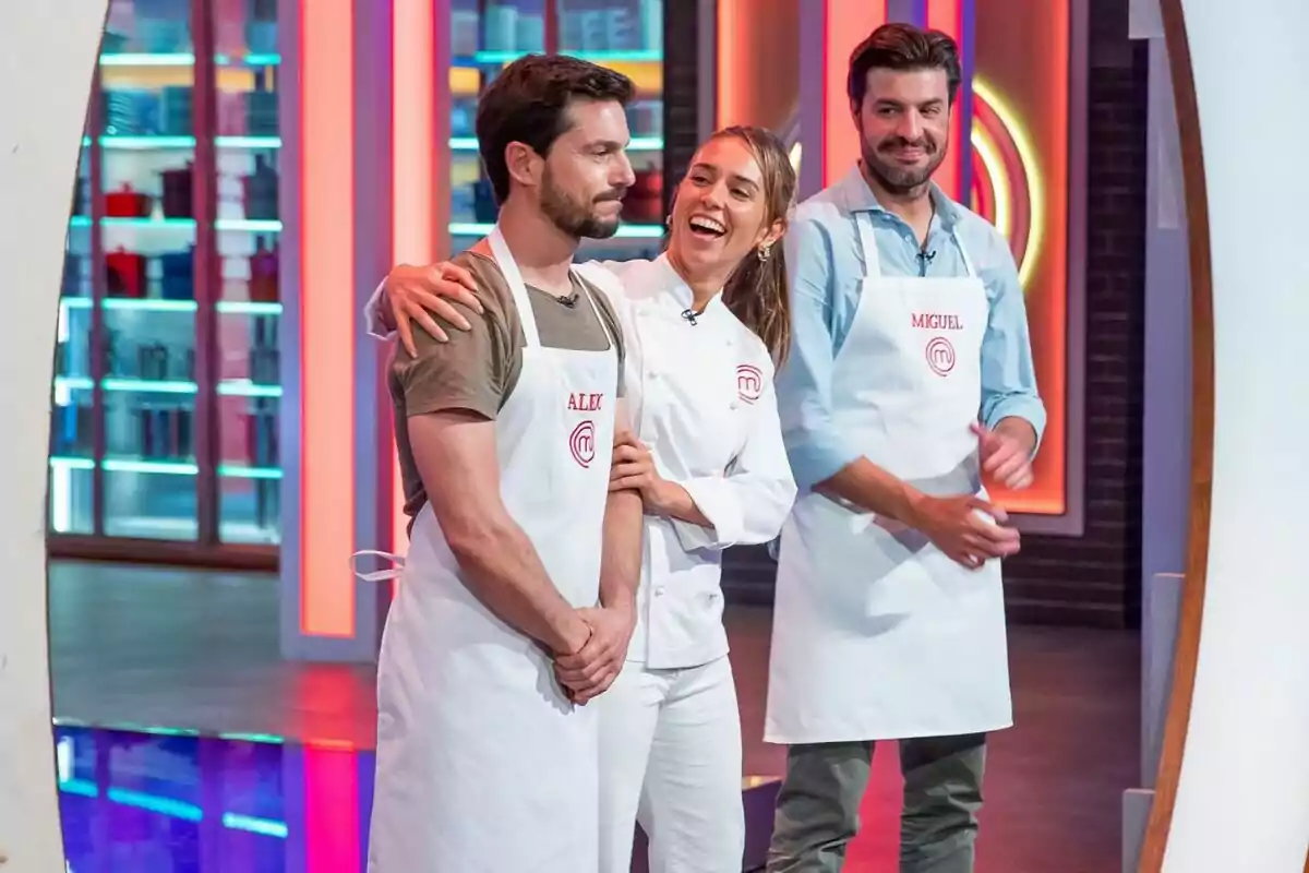 Alejo Sauras y Miguel Torres junto a una mujer con delantales de MasterChef Celebrity están en un set de cocina, una mujer sonríe y abraza a un hombre mientras otro hombre los observa sonriendo. Alejo Sauras y Miguel Torres junto a una mujer con delantales de MasterChef Celebrity están en un set de cocina, una mujer sonríe y abraza a un hombre mientras otro hombre los observa sonriendo.