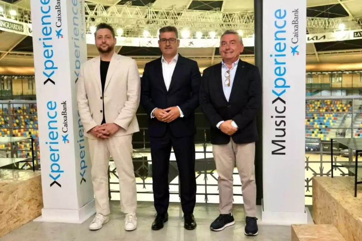 Tres hombres posan juntos en un recinto cubierto entre dos columnas blancas con el texto Xperience y Music Xperience CaixaBank, al fondo se ven gradas multicolores y una estructura metálica en el techo.