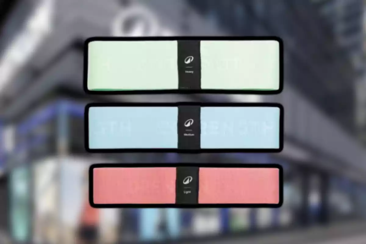 Tres bandes elàstiques de resistència de color verd, blau i rosa amb etiquetes negres al centre sobre un fons desenfocat d’edifici.