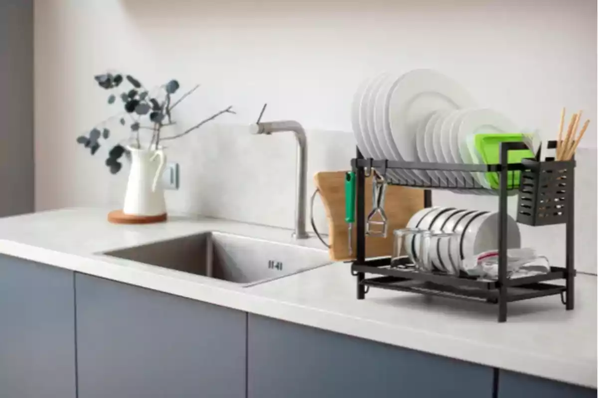 Escorreplats negre sobre una tauleta blanca al costat d’una pica amb plats, gots i utensilis organitzats; al fons, una gerra blanca amb branques decoratives. Escorreplats negre sobre una tauleta blanca al costat d’una pica amb plats, gots i utensilis organitzats; al fons, una gerra blanca amb branques decoratives.