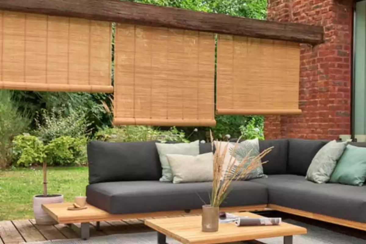 Sala exterior amb sofà en L de coixins grisos, taula de centre de fusta, persianes enrotllables de bambú i plantes al voltant