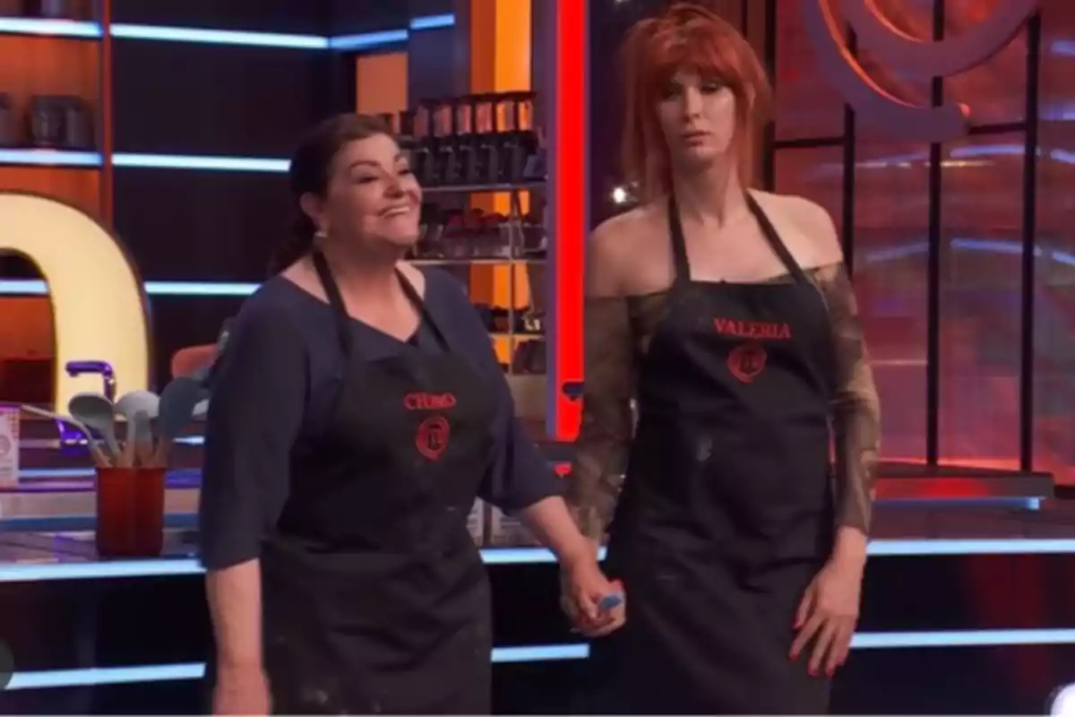 Valeria Vegas y Charo Reina en 'Masterchef Celebrity' con delantales negros que tienen sus nombres bordados, Charo sonríe mientras Valeria luce seria y ambas se toman de la mano.