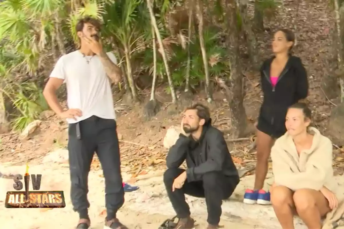 Quatre persones amb roba informal són a la platja al costat d’una zona de vegetació, dues dretes i dues assegudes, amb expressions serioses i el logotip d’un programa a la cantonada inferior esquerra.