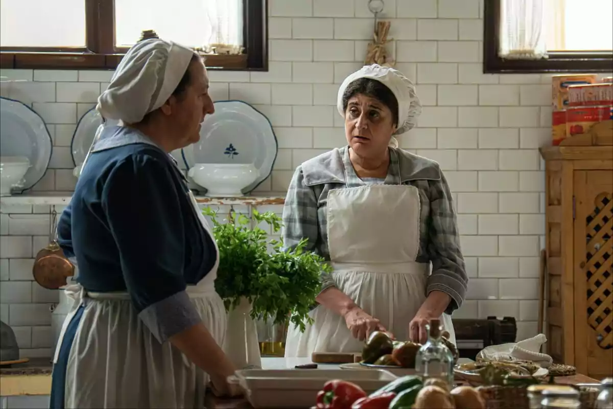 Dues dones amb davantal i còfia conversen en una cuina antiga mentre preparen aliments sobre una taula. Dues dones amb davantal i còfia conversen en una cuina antiga mentre preparen aliments sobre una taula.