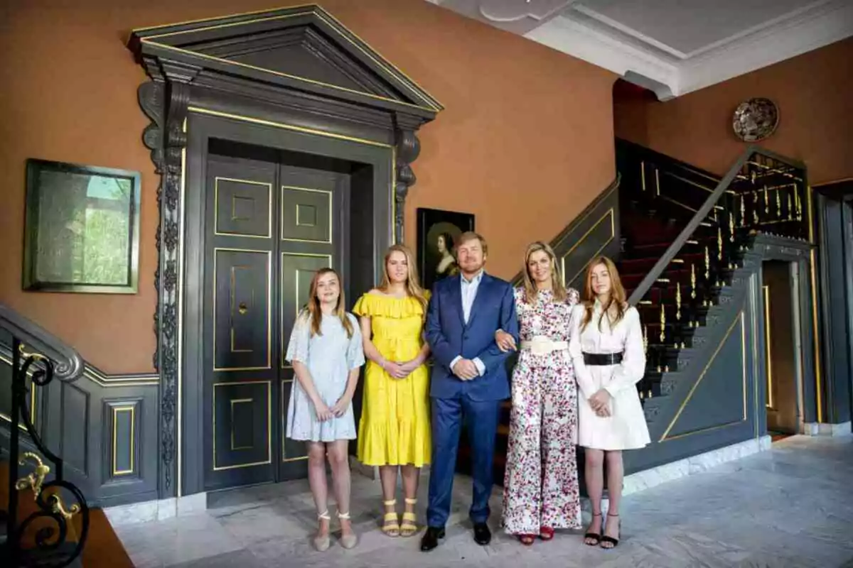 The Royal Family from the Netherlands in the foyer of an elegant house with brown walls, a large door, and a decorated staircase in the background.