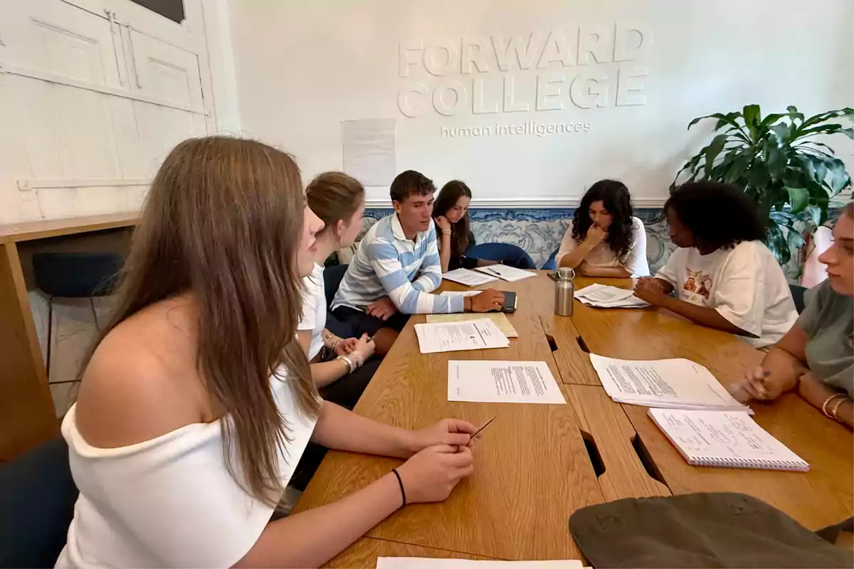 Un grup de joves asseguts, entre ells la infanta Sofia, al voltant d’una taula de fusta en una sala d’estudi, conversant i revisant documents, amb el logotip de Forward College a la paret del fons.