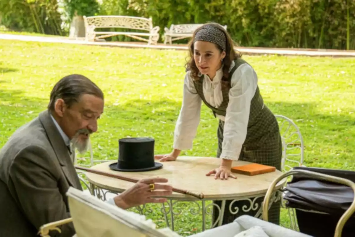 Un home gran i una dona jove conversen en un jardí asseguts en una taula rodona de ferro blanc.