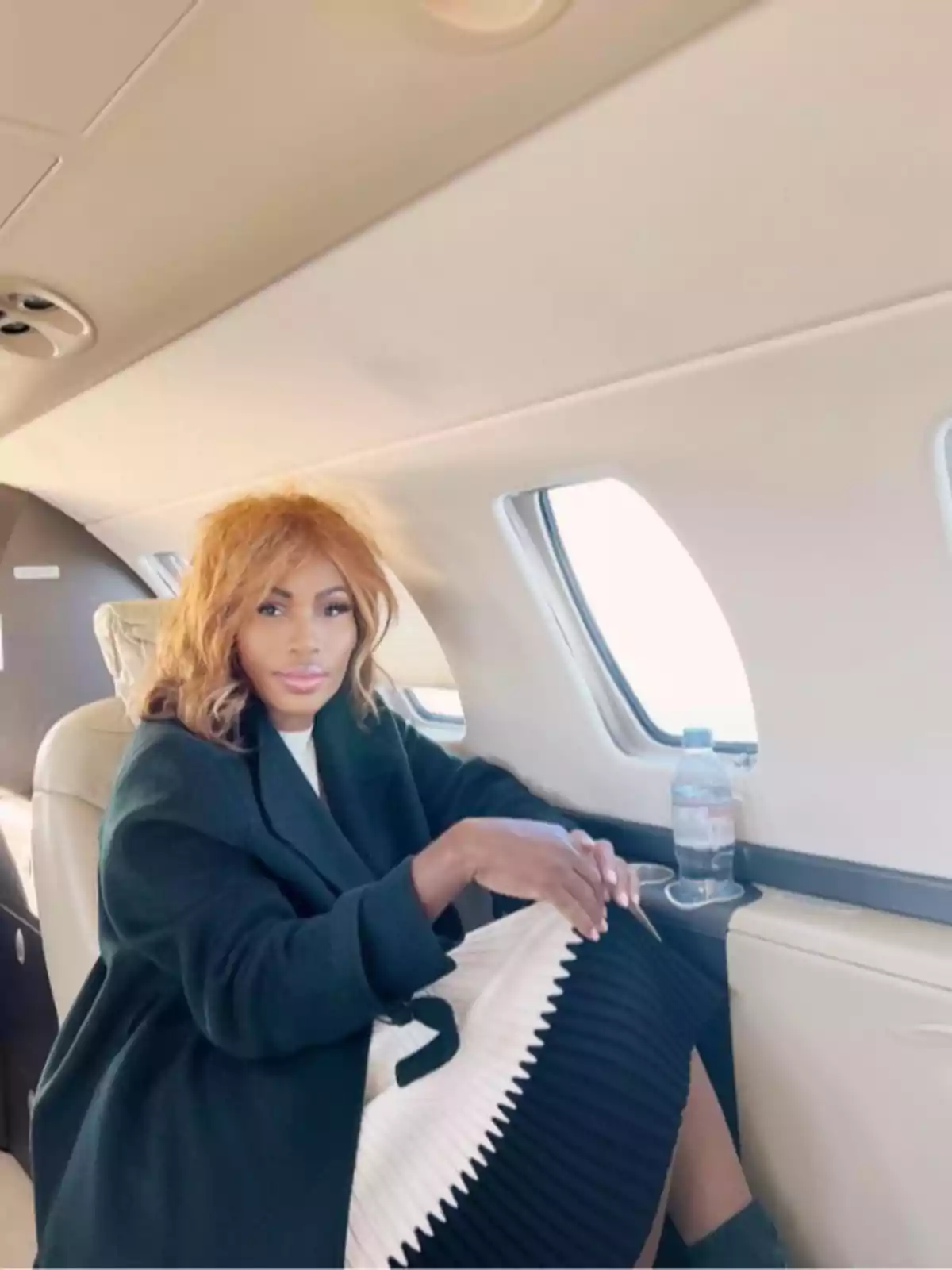 Nicole Coste sentada junto a la ventana de un avión privado vestida con abrigo oscuro y falda blanca y negra