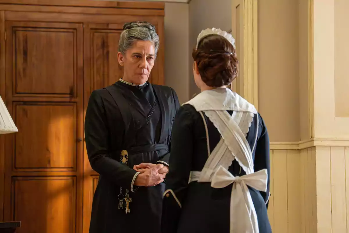 Dues dones vestides amb uniformes d’època, una d’elles amb claus penjades al cinturó, conversen a l’interior d’una casa antiga. Dues dones vestides amb uniformes d’època, una d’elles amb claus penjades al cinturó, conversen a l’interior d’una casa antiga.