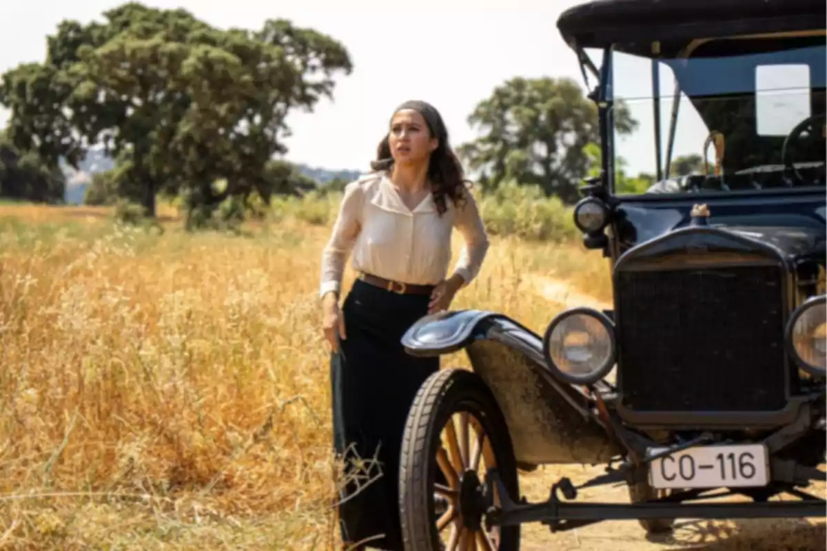 Dona vestida amb roba antiga dreta al costat d’un cotxe clàssic en un camí rural envoltat d’herba seca i arbres al fons