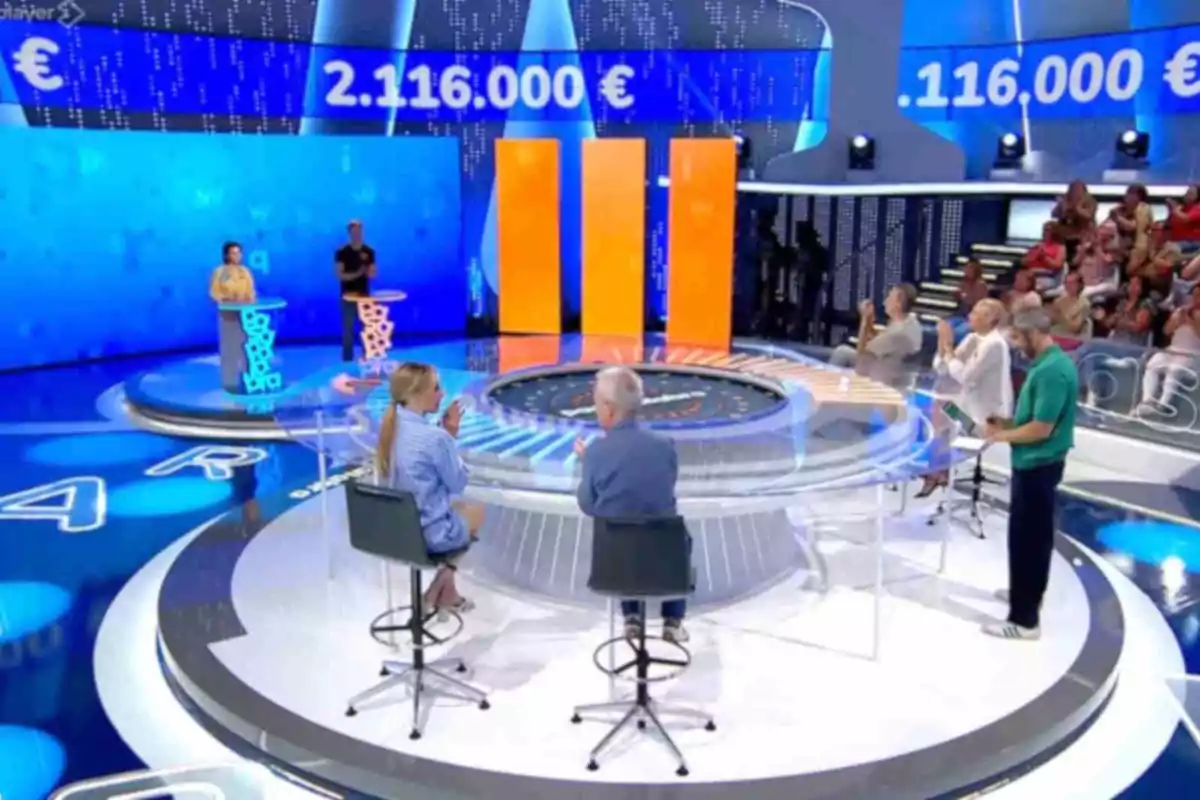 Un plató de concurs televisiu amb participants, presentador i públic, on es mostren grans xifres de diners en una pantalla blava de fons.