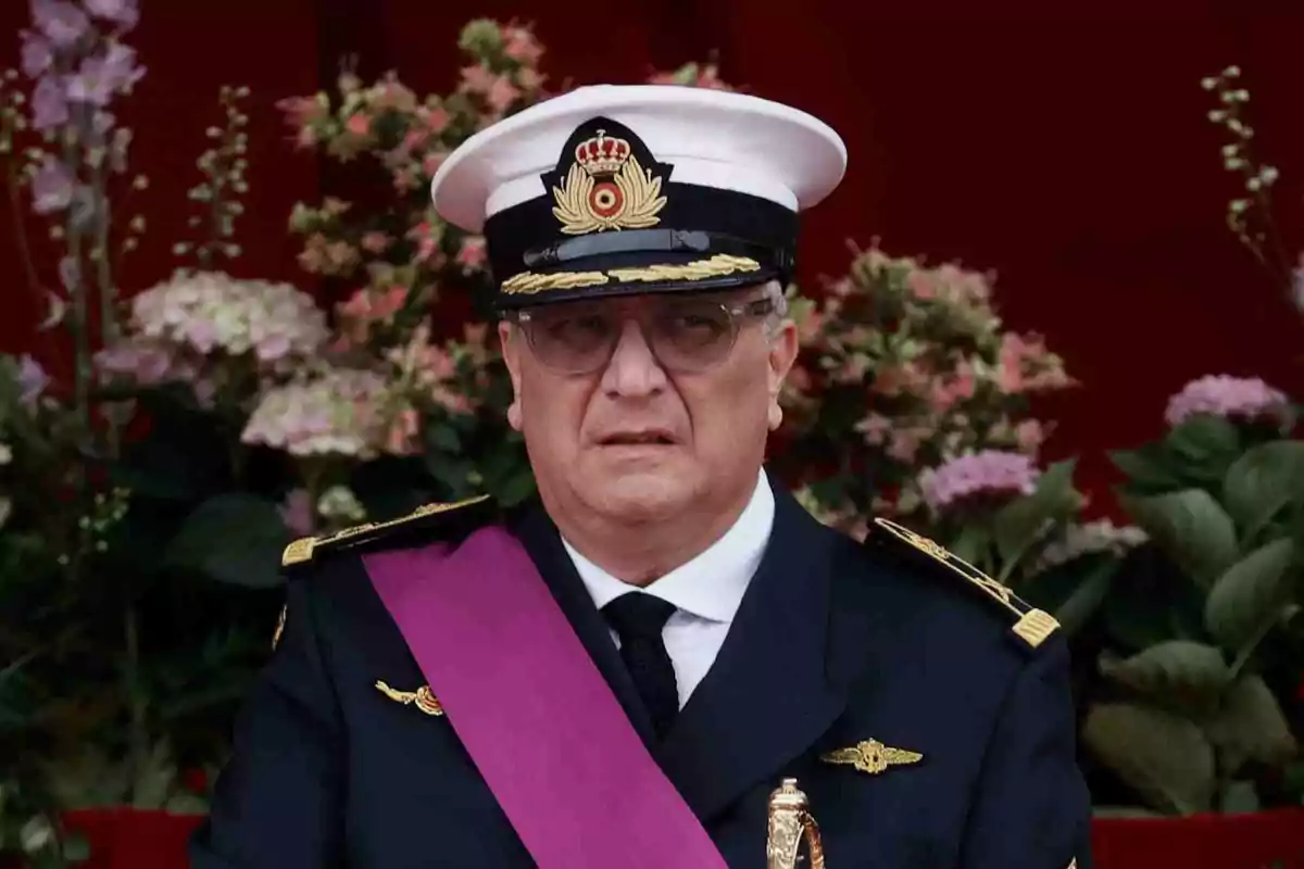 El príncipe Lorenzo de Bélgica con uniforme militar de gala, gorra blanca y banda morada, posando frente a un fondo de flores.