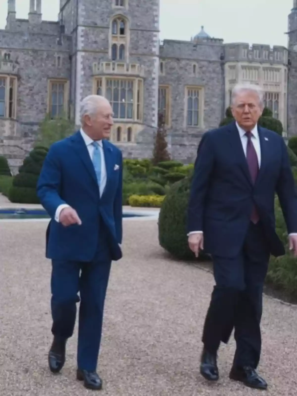 Donald Trump i el rei Carles III amb vestit caminant i conversant en un jardí davant d’un edifici antic de pedra