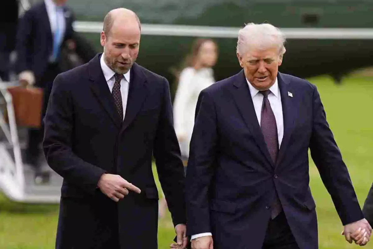 El príncep Guillem i Donald Trump vestits amb vestit fosc caminen junts sobre la gespa mentre conversen, al fons s’observa una persona desenfocada i part d’un helicòpter. El príncep Guillem i Donald Trump vestits amb vestit fosc caminen junts sobre la gespa mentre conversen, al fons s’observa una persona desenfocada i part d’un helicòpter.