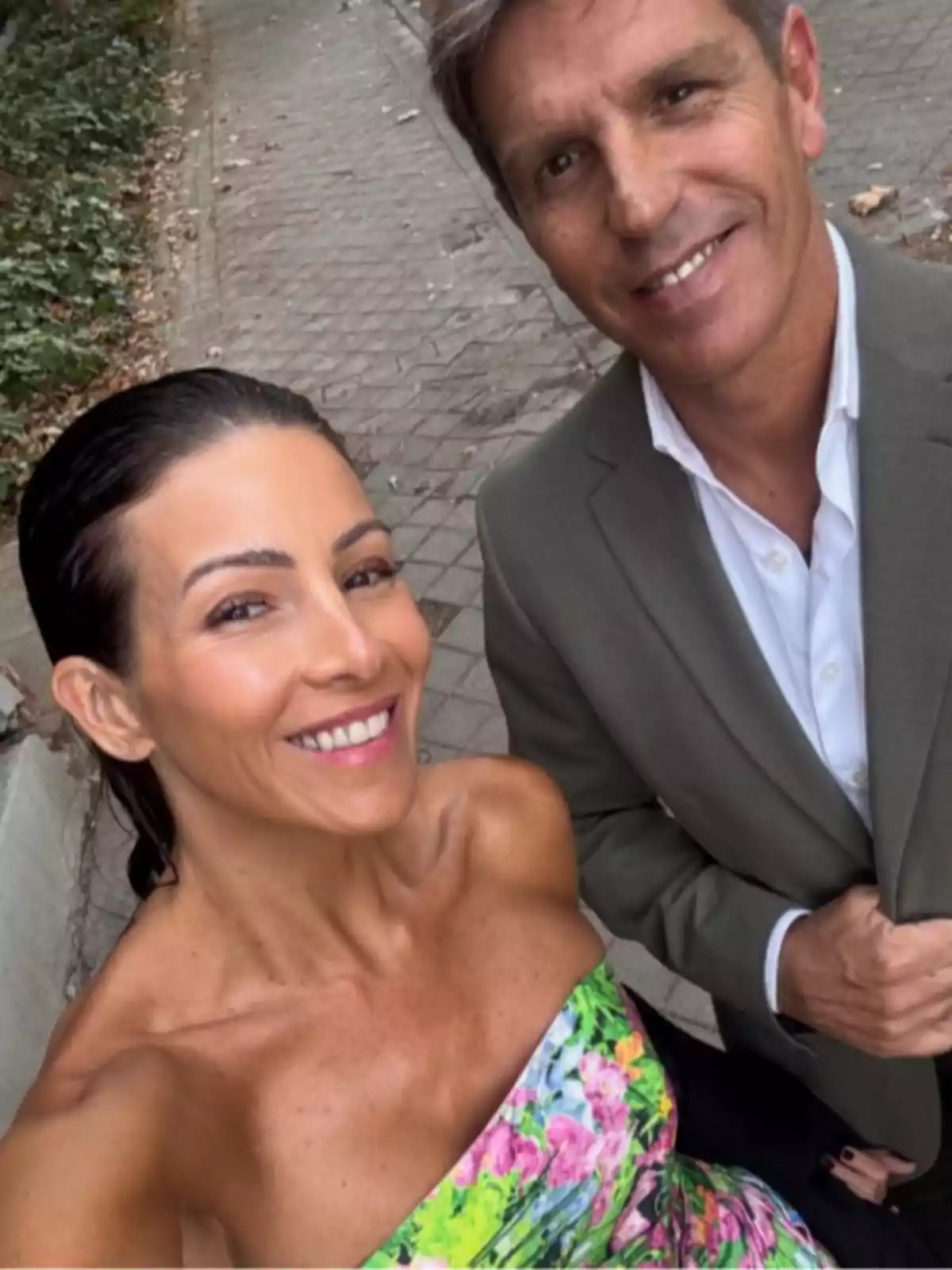 Virginia Troconis con vestido colorido y Manuel Díaz 'El Cordobés' con saco gris sonríen mientras se toman una selfie al aire libre sobre una acera.