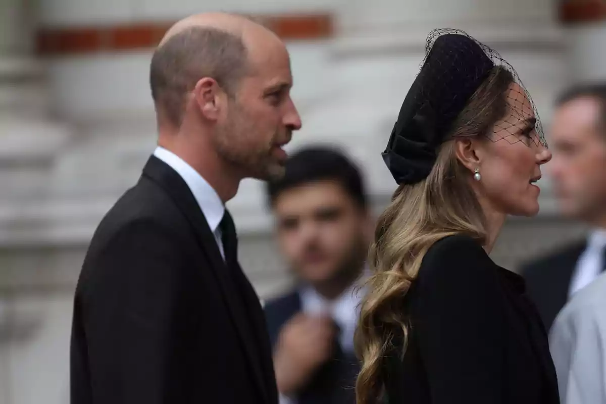 Kate Middleton i el príncep Guillem vestits de negre caminen un al costat de l’altra en un esdeveniment formal; Kate porta un tocat negre amb vel i arracades de perla. Kate Middleton i el príncep Guillem vestits de negre caminen un al costat de l’altra en un esdeveniment formal; Kate porta un tocat negre amb vel i arracades de perla.