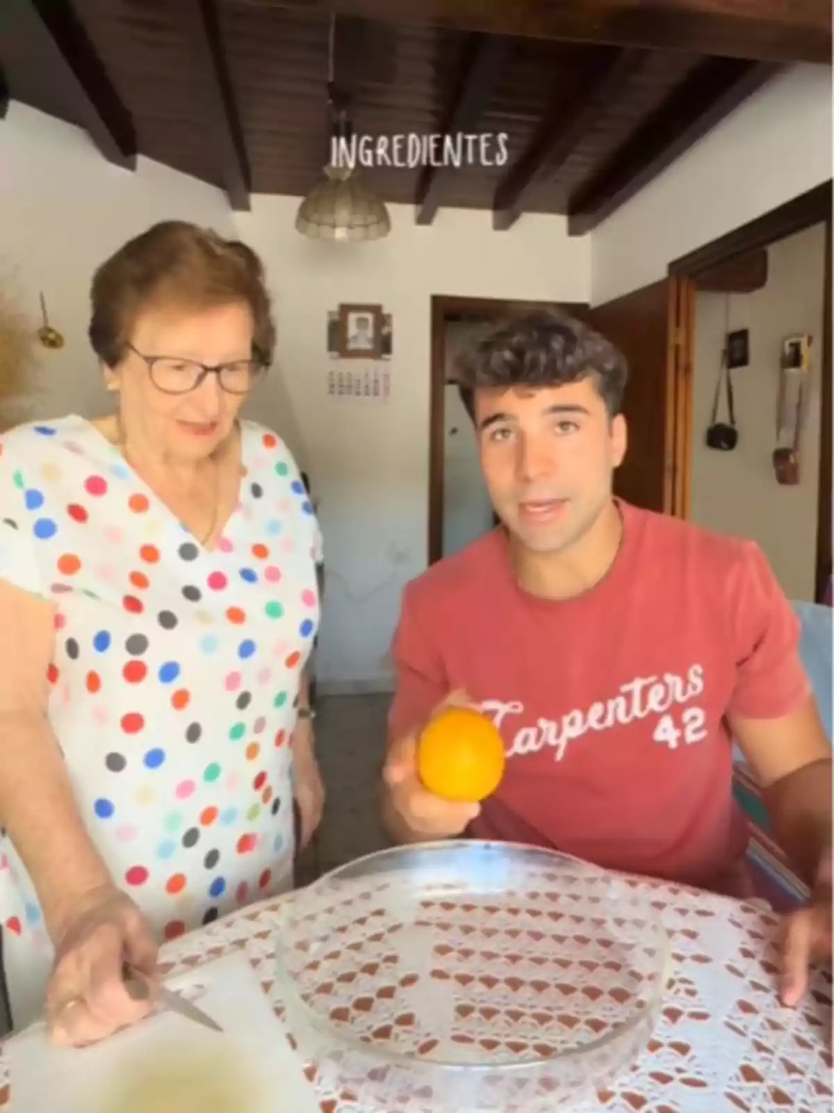 Una dona gran i un home jove són a una cuina, l’home sosté una taronja i la dona un ganivet, damunt d’una taula amb estovalla de puntes i una safata de vidre. Una dona gran i un home jove són a una cuina, l’home sosté una taronja i la dona un ganivet, damunt d’una taula amb estovalla de puntes i una safata de vidre.