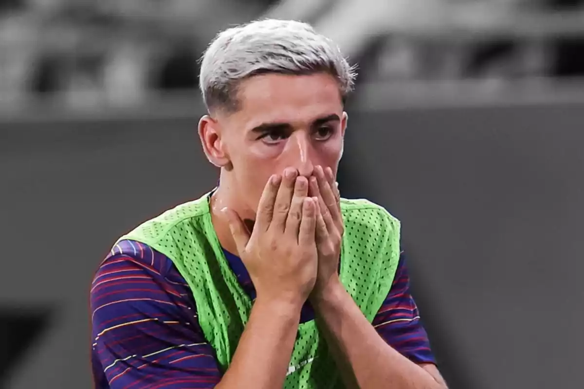 Jove futbolista amb els cabells tenyits de ros cobrint-se la boca amb totes dues mans i amb una expressió de sorpresa o preocupació Jove futbolista amb els cabells tenyits de ros cobrint-se la boca amb totes dues mans i amb una expressió de sorpresa o preocupació