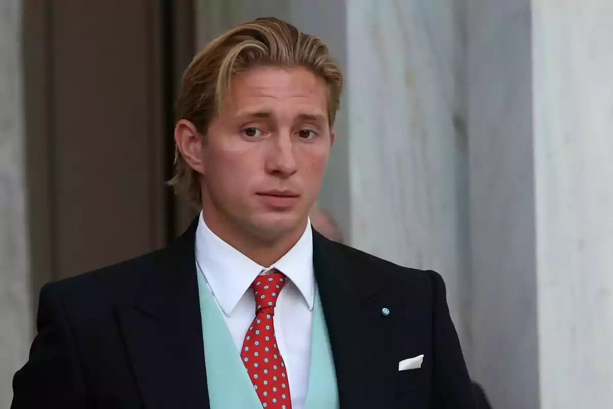 Home jove de cabell ros pentinat cap enrere vestit amb un vestit fosc, armilla blava cel i corbata vermella amb punts blaus davant d’una paret de marbre blanc Home jove de cabell ros pentinat cap enrere vestit amb un vestit fosc, armilla blava cel i corbata vermella amb punts blaus davant d’una paret de marbre blanc