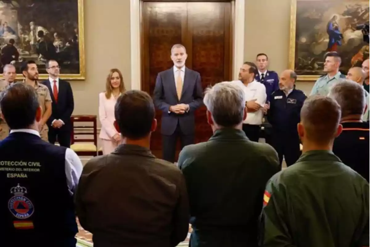 Un grup de persones amb uniformes i vestits formals escolta un home amb vestit que parla en una sala elegant amb quadres grans a les parets. Un grup de persones amb uniformes i vestits formals escolta un home amb vestit que parla en una sala elegant amb quadres grans a les parets.