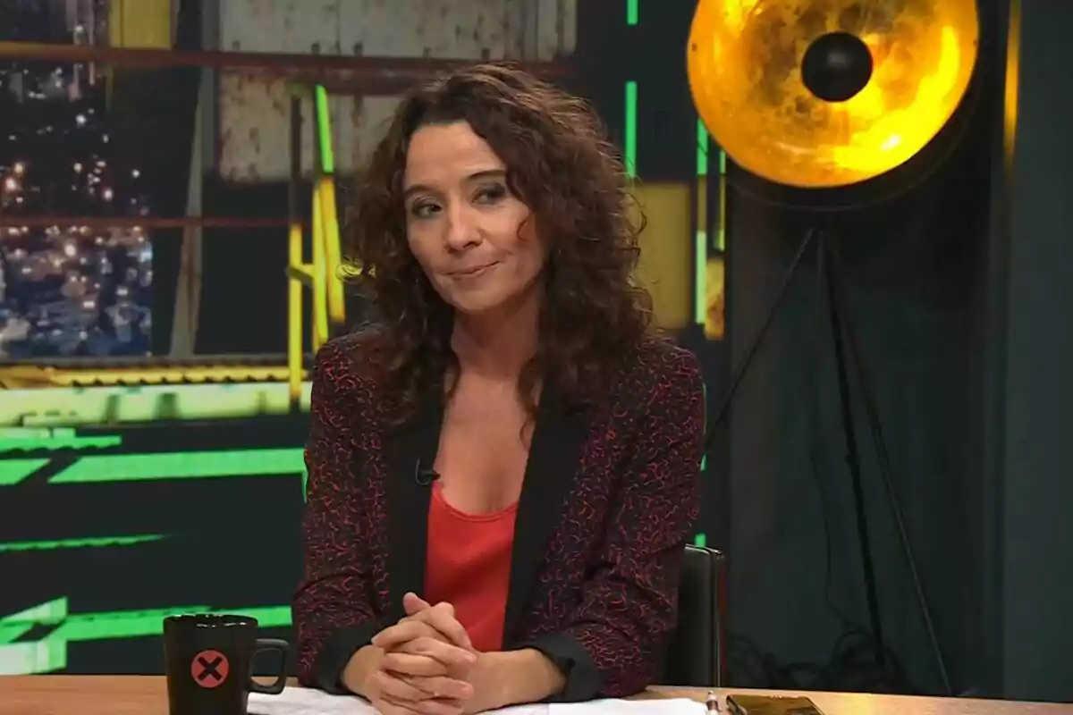 Dona de cabells arrissats i foscos asseguda en un estudi de televisió amb una tassa negra sobre la taula i una llum groga de fons Dona de cabells arrissats i foscos asseguda en un estudi de televisió amb una tassa negra sobre la taula i una llum groga de fons