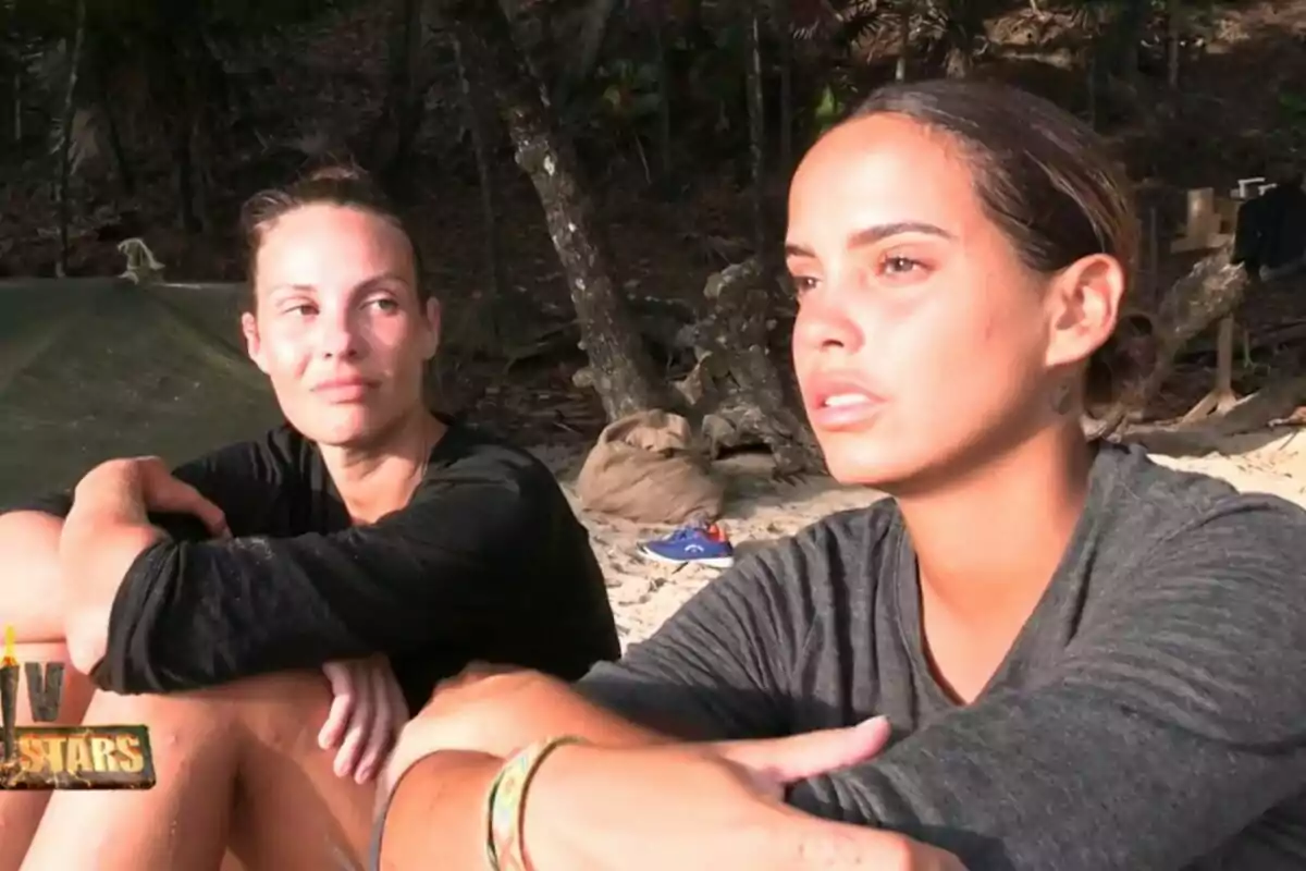 Dues dones assegudes a la sorra d’una platja amb expressió seriosa envoltades d’arbres i vegetació