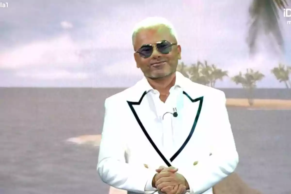 Home amb ulleres de sol i vestit blanc posant davant d’un fons de platja amb palmeres