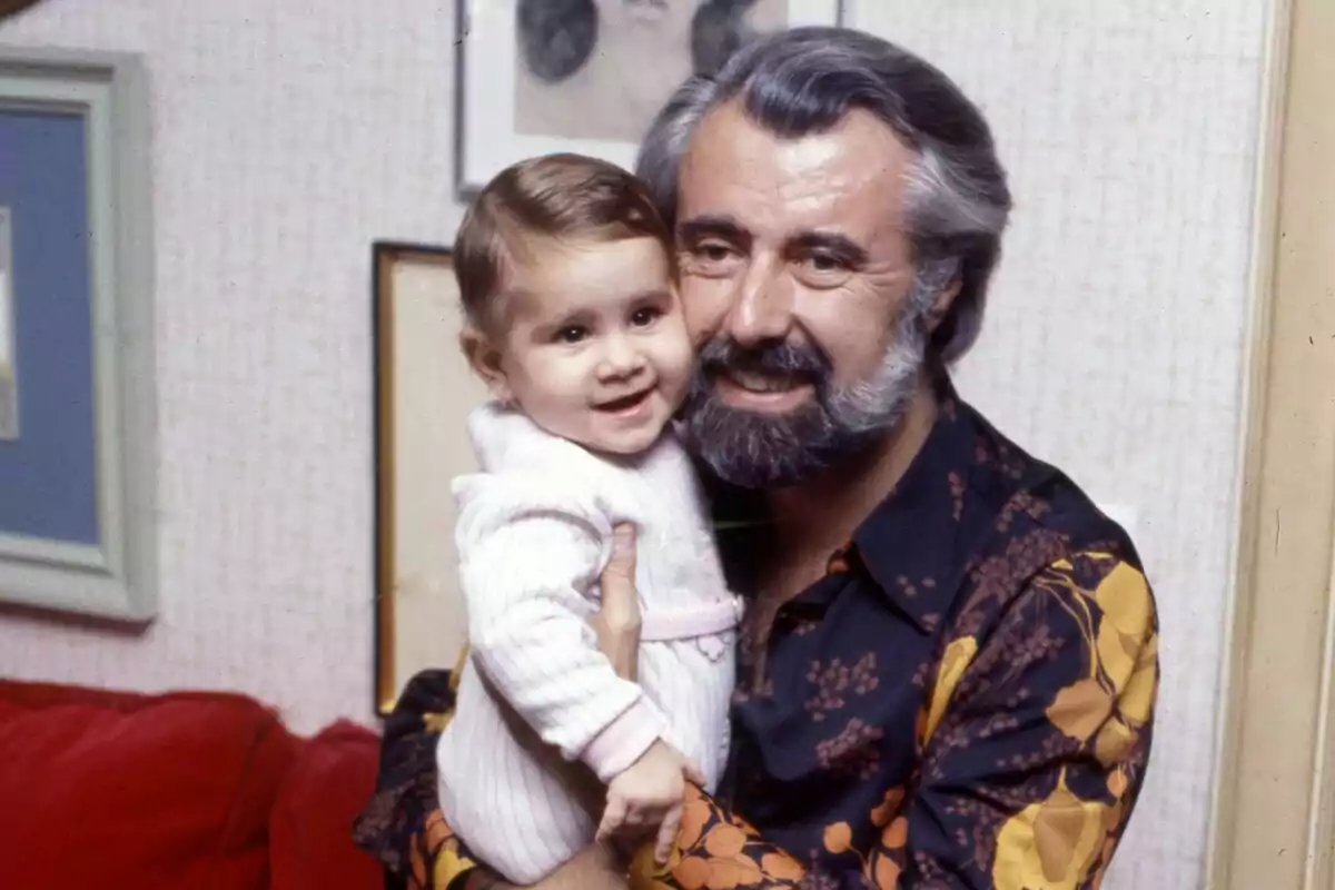 Home de cabells canosos i barba sosté en braços una nadó somrient en una sala decorada amb quadres.