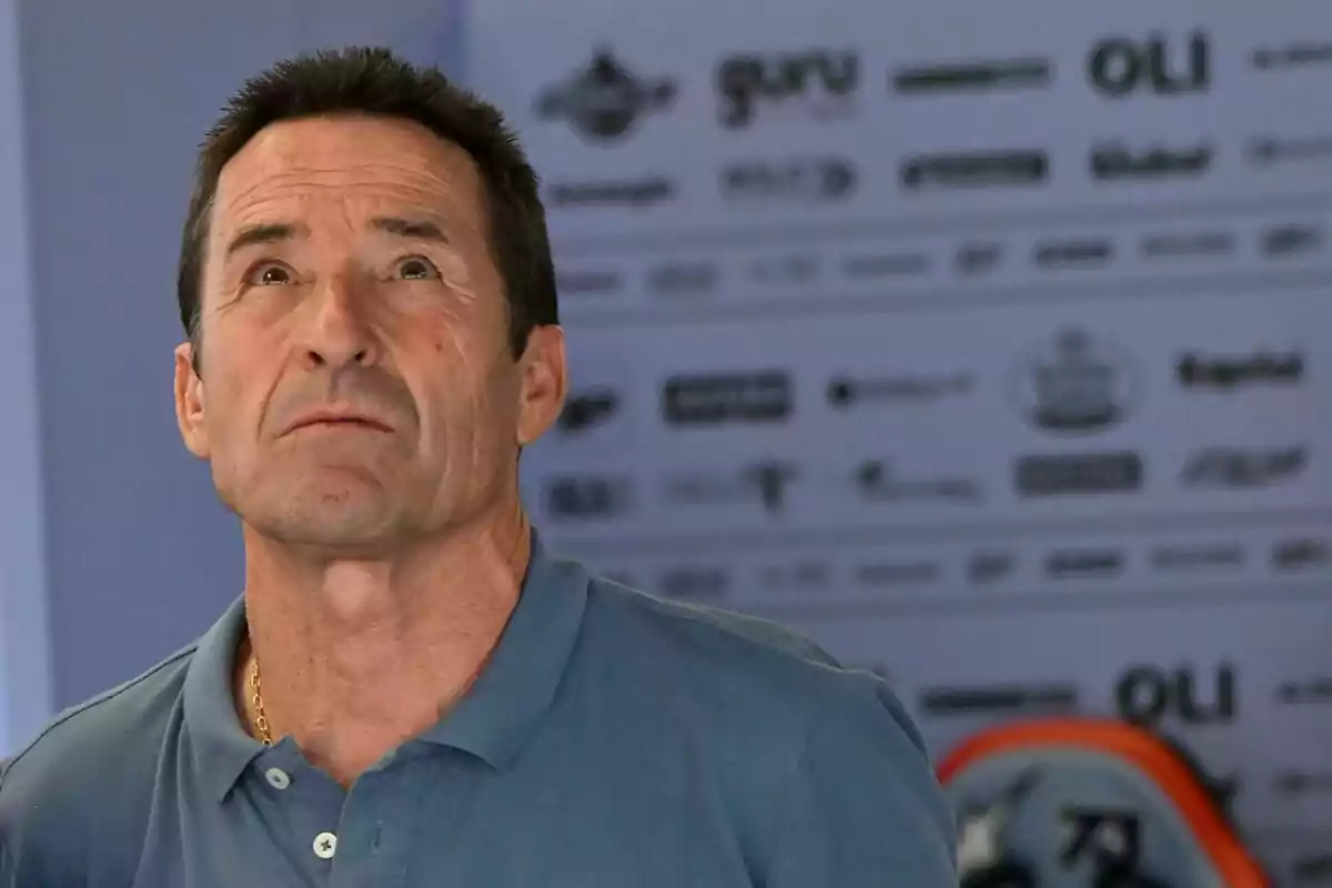 Hombre de cabello corto y oscuro con camisa tipo polo azul mirando hacia arriba con expresión seria y fondo desenfocado con logotipos. Hombre de cabello corto y oscuro con camisa tipo polo azul mirando hacia arriba con expresión seria y fondo desenfocado con logotipos.