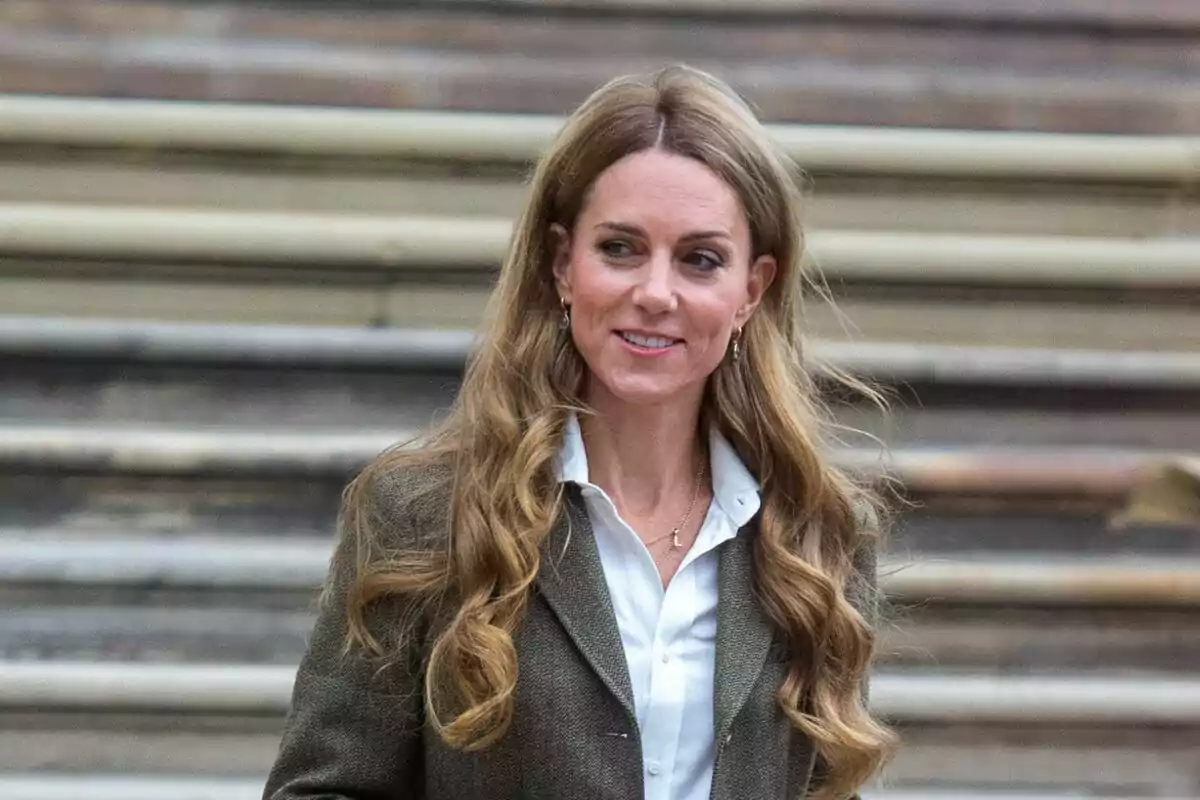 Dona de cabell llarg i ondulat amb jaqueta marró i camisa blanca dreta davant d’unes escales Dona de cabell llarg i ondulat amb jaqueta marró i camisa blanca dreta davant d’unes escales