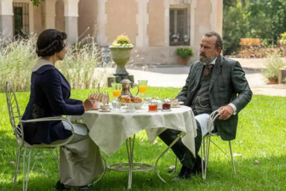Dues persones vestides amb roba d’època estan assegudes en un jardí esmorzant en una taula rodona coberta amb un mantel blanc.