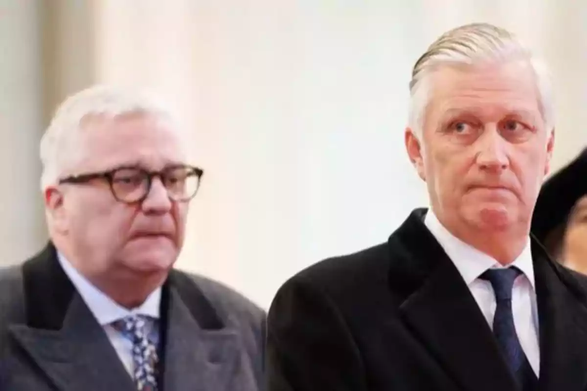 Dos homes grans vestits amb vestit formal miren cap al davant amb expressions serioses en un entorn interior il·luminat. Dos homes grans vestits amb vestit formal miren cap al davant amb expressions serioses en un entorn interior il·luminat.