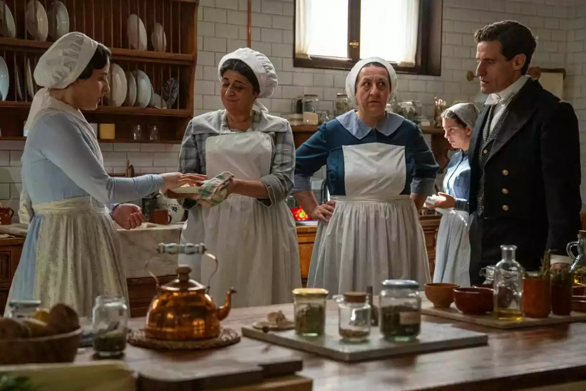 Cinc persones vestides amb roba d’època conversen en una cuina antiga mentre preparen menjar envoltades d’estris i pots sobre una taula de fusta. Cinc persones vestides amb roba d’època conversen en una cuina antiga mentre preparen menjar envoltades d’estris i pots sobre una taula de fusta.