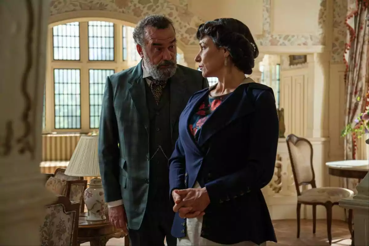 Un home i una dona vestits de manera elegant conversen en una sala decorada amb mobles clàssics i grans finestres al fons.