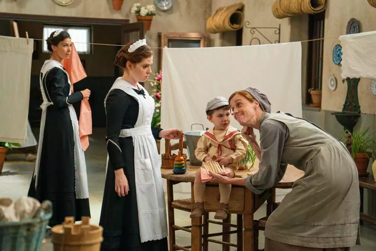 Tres dones vestides de minyones i un nen petit amb roba antiga són en una bugaderia, envoltats de roba estesa i estris de neteja, mentre una de les dones interactua de manera afectuosa amb el nen assegut damunt d’una taula.